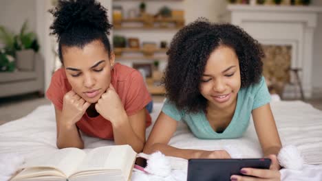 Two-cheerful-mixed-race-funny-girlfriends-reading-book-and-using-tablet-computer-talks-and-have-fun-lying-in-bed-at-home