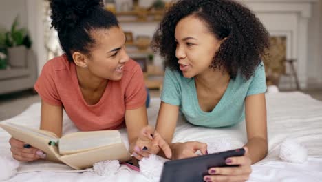 Cheerful-mixed-race-funny-girl-using-tablet-computer-show-photos-on-tablet-computer-to-her-sister-reading-book-and-lying-in-bed-at-home