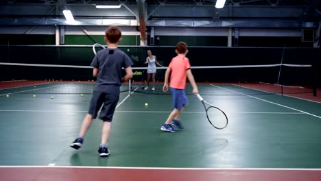 A-female-trainer-in-a-sports-suit-teaches-playing-tennis-in-two-middle-aged-boys,-guys-are-bouncing-balls-on-a-tennis-court