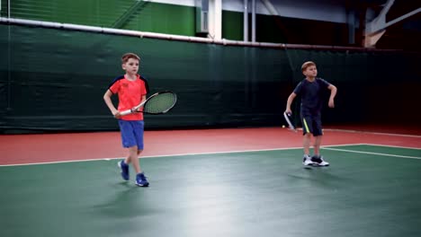 Two-young-boys-with-rackets-playing-tennis-on-court-together