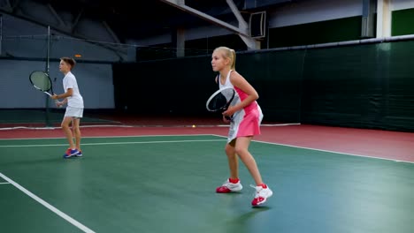 Two-young-athletes-in-recreation-area-playing-sport-game.-Happy-sister-and-brother-having-tennis-lesson-spending-time-at-indoor-court.-Girl-and-boy-serving-and-returning-yellow-balls-with-rackets