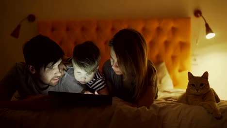 Happy-family-with-little-son-and-funny-cat-lying-in-bed-at-home-and-surfing-social-media-on-tablet-computer-before-sleeping