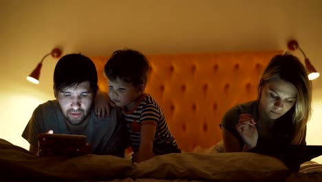 Familia-feliz-con-pequeño-hijo-tumbado-en-la-cama-en-el-hogar-usando-computadoras-tablet-y-smartphone-para-ver-la-película-y-dibujos-animados-en-la-noche-antes-de-dormir