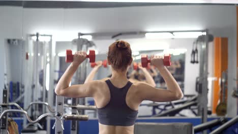 Joven-hermosa-chica-deportiva-haciendo-press-de-hombros-con-pesas-ejercicio-en-un-gimnasio-de-deporte,-vista-parte-trasera