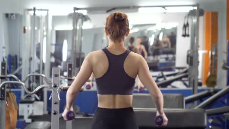 Sports-young-girl-doing-side-dumbbell-raises-while-sitting-on-sports-bench-in-a-gym.-Back-side-view-60-fps-Close-up