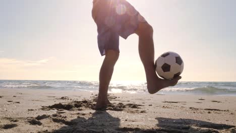 Hombre-joven-en-bañador-haciendo-trucos-con-fútbol-en-el-pie-en-la-orilla-del-mar-en-la-playa-de-silueta-al-atardecer