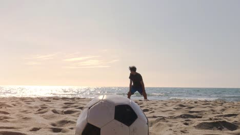 Young-man-goalkeeper-in-swimming-trunks-saves-football-penalty-on-the-sea-shore-at-the-beach-at-sunset-silhouette