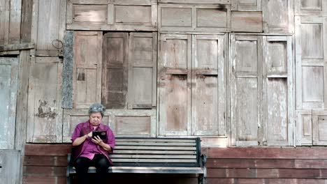 Asia-mujer-mayor-sosteniendo-el-teléfono-móvil-al-lado-de-la-vieja-casa-de-madera.-mensaje-de-texto-mujeres-mayores,-con-la-aplicación-de-teléfono-inteligente-en-el-parque.-teléfono-móvil-de-uso-principal-para-conectar-con-la-red-social