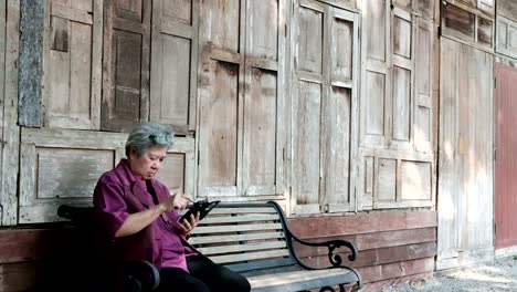 asian-elder-woman-holding-mobile-phone-beside-old-wood-house.-elderly-female-texting-message,-using-app-with-smartphone-in-park.-senior-use-cellphone-to-connect-with-social-network