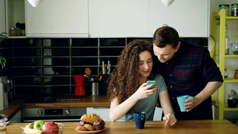 mujer-caucásica-feliz-rizada-atractiva-joven-está-sentado-en-mensajes-de-texto-de-cocina-alguien-via-smartphone,-viene-de-su-marido-y-ella-muestra-algo-gracioso-en-el-teléfono