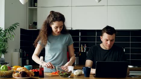 Cheerful-couple-at-table-woman-dancing-and-cooking-while-gives-hasband-to-try-red-pepper,-man-sitting-working-on-laptop,-he-shows-something-funny-on-screen