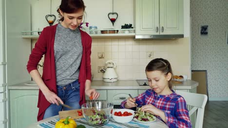 Happy-young-mother-and-cheerful-daughter-cook-salad-together-in-the-kitchen-at-home-cutting-vegetables-and-talking.-Family,-cooking,-and-people-concept