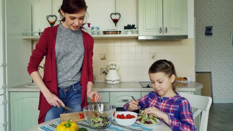 Glückliche-junge-Mutter-und-fröhliche-Tochter-Kochen-Salat-zusammen-in-der-Küche-zu-Hause-Schneiden-von-Gemüse-und-sprechen.-Familie,-Kochen-und-Menschen-Konzept