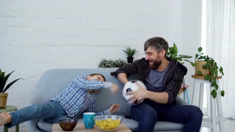 Adorable-little-kid-is-playing-with-ball-with-his-active-caring-father,-throwing-and-catching-football-on-couch-at-home.-Family-bonding-and-funny-sport-concept.
