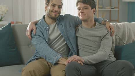 Close-Up-Shot-of-a-Cute-Male-Gay-Couple-Sitting-Together-on-a-Sofa-at-Home.-They-are-Hugging-and-Holding-Hands.-They-are-Happy-and-Smiling.-They-are-Casually-Dressed-and-Room-Has-Modern-Interior.