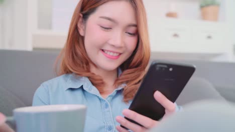 Portrait-of-beautiful-attractive-smiling-Asian-woman-using-smartphone-holding-a-warm-cup-of-coffee-or-tea-while-lying-on-the-sofa-when-relax-in-living-room-at-home.-Lifestyle-women-at-home-concept.