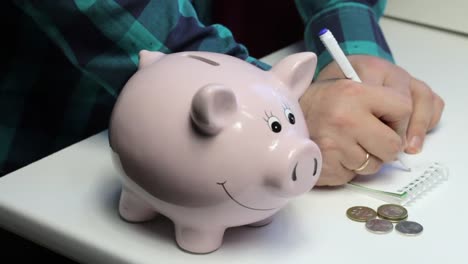 On-the-table-there-is-a-piggy-bank-in-the-form-of-a-pink-pig.-The-man-in-the-hands-of-the-coin,-which-he-puts-in-turn-in-a-piggy-bank.-Makes-notes-in-a-notebook.