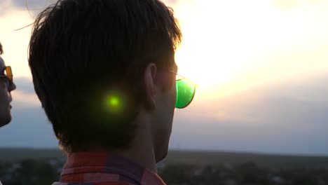 Close-up-of-young-male-couple-in-sunglasses-standing-on-the-edge-of-rooftop-and-talking.-Handsome-gay-boys-rest-on-roof-of-high-rise-building-and-enjoying-beautiful-cityscape.-Dolly-shot-Slow-motion