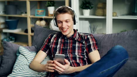 El-joven-feliz-en-los-auriculares-está-escuchando-música-y-usando-el-teléfono-inteligente-en-casa