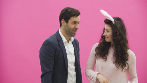 Young-couple-standing-on-a-pink-background.-The-wife-of-a-pink-rabbit-on-her-head,-holding-in-her-hand-rabbit-ears-and-looking-in-love-with-her-husband.-Happy-family-is-preparing-for-Easter.