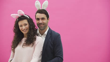 Una-joven-pareja-de-pie-sobre-un-fondo-rosa.-Durante-esto,-miran-a-la-cámara-y-muestran-un-gesto-de-llamada.-La-familia-feliz-se-está-preparando-para-la-Pascua,-con-las-orejas-de-un-conejo-rosa-en-la-cabeza.