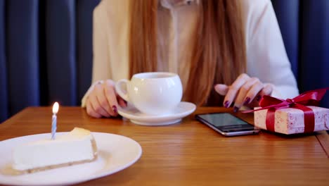 Mujer-pelirroja-irreconocible-sentada-en-la-cafetería-y-navegando-en-el-teléfono-celular-en-su-cumpleaños.-Taza-de-té,-caja-de-regalo-y-pedazo-de-pastel-con-vela-ardiente-en-su-mesa