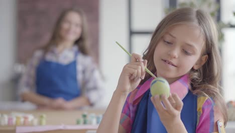 Portrait-of-cute-concentrated-girl-with-long-hair-in-blue-apron-painting-Easter-egg