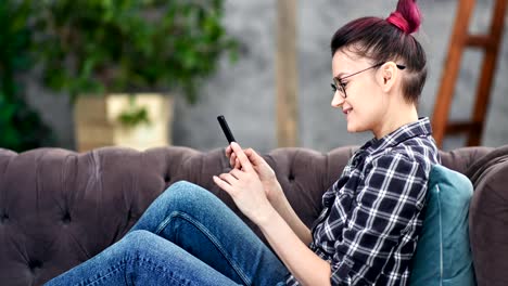 Lächelnde-Hipster-junge-Frau-plaudert-mit-Smartphone-sitzen-auf-Couch-zu-Hause-mittel-erschossen