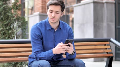 Hombre-joven-escribiendo-mensaje-en-el-teléfono-inteligente,-sentado-al-aire-libre-en-el-Banco