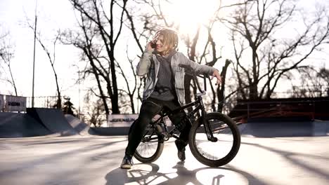 Portrait-of-happy-handsome-teenage-boy-with-dreadlocks,-using-mobile-phone-while-riding-a-BMX-bike,-smiling,-messaging-friends-via-social-networks.-People,-lifestyle-and-communication-concept.-Low-angle-footage