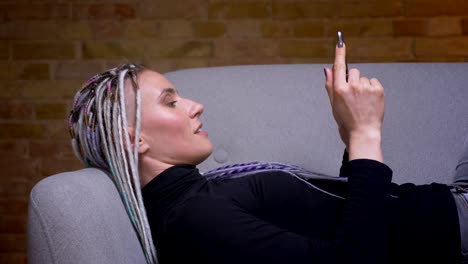 Closeup-shoot-of-young-attractive-caucasian-hipster-female-using-the-tablet-turning-to-camera-and-happily-smiling-lying-on-the-couch-indoors-in-a-cozy-apartment