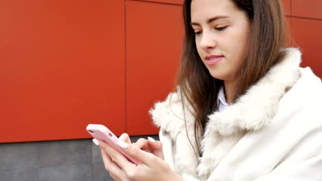 A-young-attractive-girl-stands-at-the-wall-of-a-building-and-pokes-a-finger-into-a-mobile-phone.