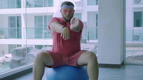 Funny-fat-male-in-pink-glasses-and-in-a-pink-t-shirt-is-engaged-on-a-fit-ball-in-the-gym-depicting-a-girl.-A-wonderful-man-listens-to-music-and-dances-on-the-ball-in-the-gym