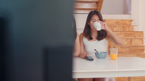 Pareja-lesbiana-asiática-viendo-la-televisión-mientras-bebe-jugo-de-bebida,-cereales-de-copos-de-maíz-y-leche-en-un-tazón-en-la-mesa-en-la-cocina-en-casa.