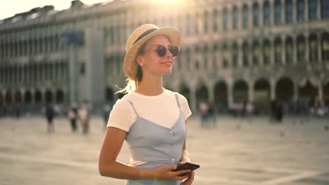 Caucasian-young-woman-tourist-in-blue-dress-walking-on-square-in-Rome-checking-mail-from-friend-on-smartphone-connected-to-4G-in-roaming