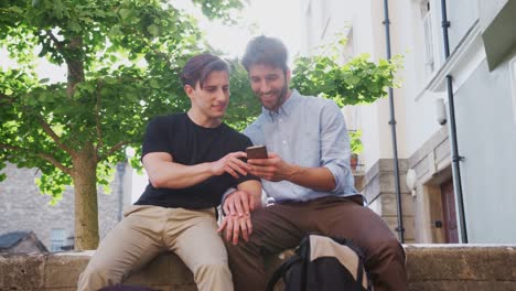 Pareja-gay-masculina-sentada-al-aire-libre-en-la-pared-de-la-construcción-mirando-el-teléfono-móvil