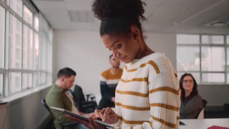 Lächelnde-junge-afrikanische-Geschäftsfrau-mit-digitalem-Tablet,-während-Kollegen-im-Hintergrund-im-Büro-interagieren