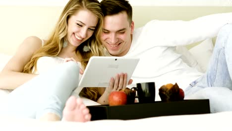 Happy-couple-watching-a-funny-movie-on-tablet-computer-together-in-bedroom.-4K