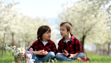 Two-children,-boy-brothers,-eating-chocolate-bunnies-and-having-fun-with-easter-eggs-in-the-park,-beautiful-spring-blooming-garden