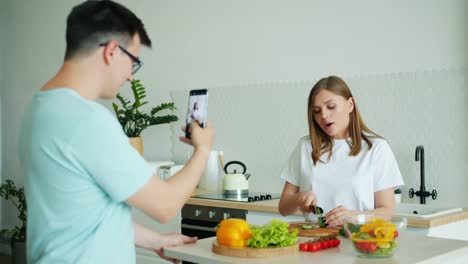 Cámara-lenta-de-la-joven-cocinando-ensalada-mientras-el-hombre-toma-fotos-con-el-teléfono-inteligente