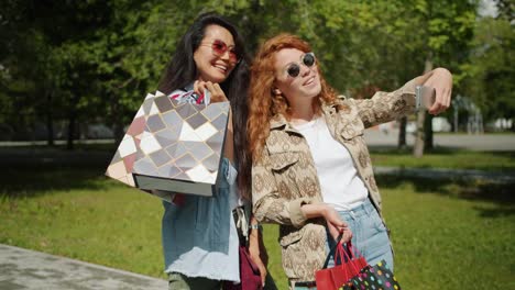 Pareja-de-chicas-tomando-selfie-con-teléfono-inteligente-sosteniendo-bolsas-de-compras-en-el-parque