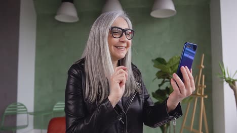 Close-up-of-beautiful-gray-hair-smiling-woman-in-black-jacket-waving-her-hand-to-the-interlocutor-while-having-video-call-at-home