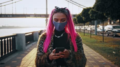 Young-hipster-woman-in-informal-clothes-and-protective-mask-is-typing-on-cellphone,-walking-by-embankment