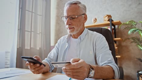 Un-hombre-de-negocios-maduro-está-sosteniendo-tarjeta-de-crédito-e-ingresando-su-número-en-el-teléfono-celular,-comprando-en-línea-sentado-en-la-mesa