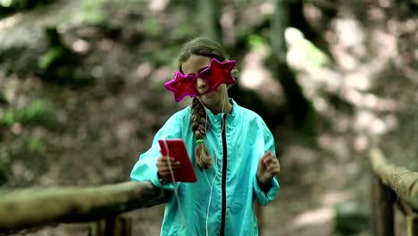 Girl-with-red-smartphone-stands-on-wooden-bridge,-listens-to-music-and-dance