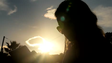 mujer-joven-con-gafas-de-sol-con-tableta,-sol-tiro,-nubes-y-silueta-de-las-palmeras