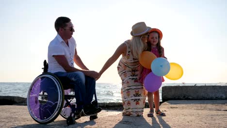 happy-daughter-sitting-on-handicapped-dad-in-wheelchair-with-balloons-in-hand,-parents-with-child-hugging