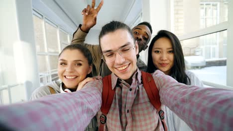 Point-of-view-of-four-positive-cheerful-attractive-multi-ethnic-friends-talking-selfie-photos-holding-smartphone-and-having-fun-while-standing-in-corridor-of-university