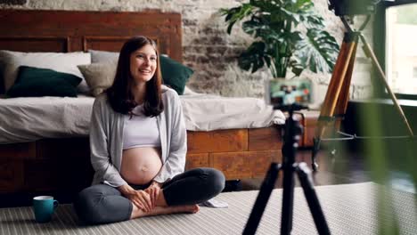 Schöne-junge-Frau-Blogger-ist-Video-über-Schwangerschaft-für-ihr-Vlog-zu-Hause-auf-dem-Boden-sitzen,-reden-und-Blick-in-die-Smartphone-Kamera-auf-Stativ-aufgenommen.