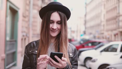 Lächelnde-junge-Europäerin-in-stilvollen-Hut-entlang-Stadtstraße-mit-einem-Smartphone-Zeitlupe-vorwärts-schauen